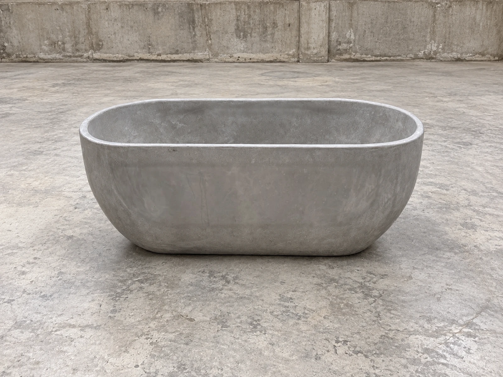 Tina ovalada de concreto gris natural con borde redondeado, sobre piso de cemento pulido frente a un muro de concreto