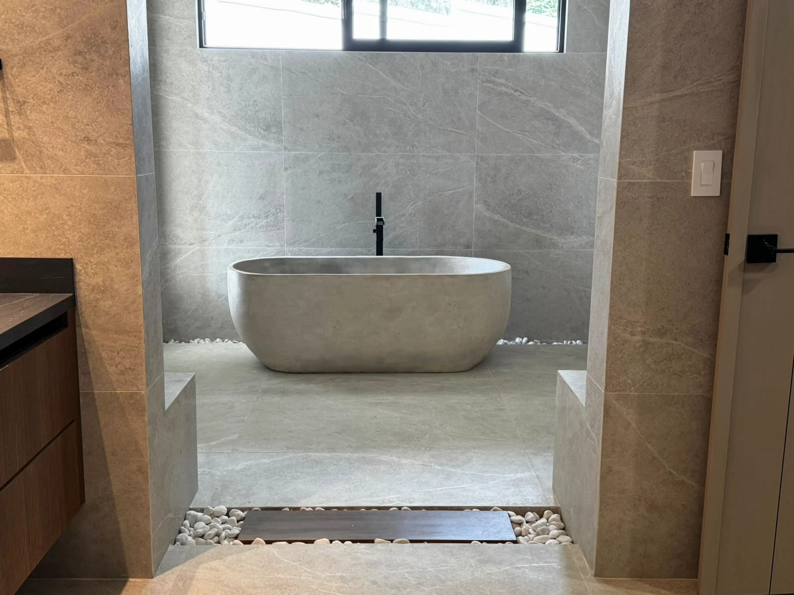 Tina ovalada de concreto gris claro al fondo de un baño con porcelanato gris, grifería negra y piedras de río en el piso
