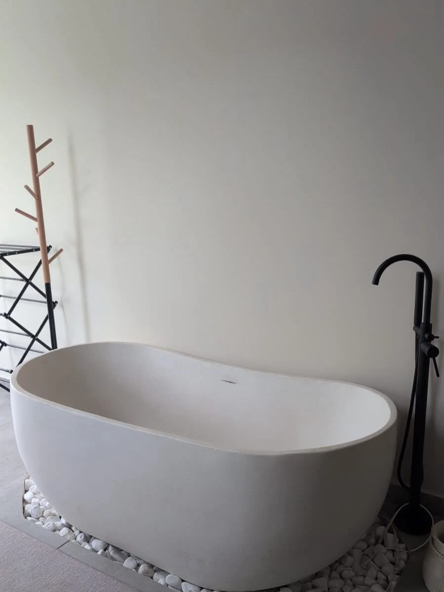 Tina ovalada de concreto blanco liso con grifería negra de piso, en baño con pared blanca y piedras decorativas en el borde