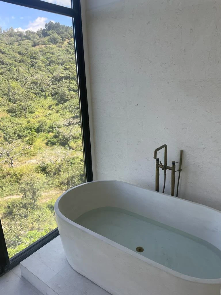 Tina ovalada de concreto blanco sobre una plataforma, junto a un ventanal alto con vista al bosque y grifería de piso