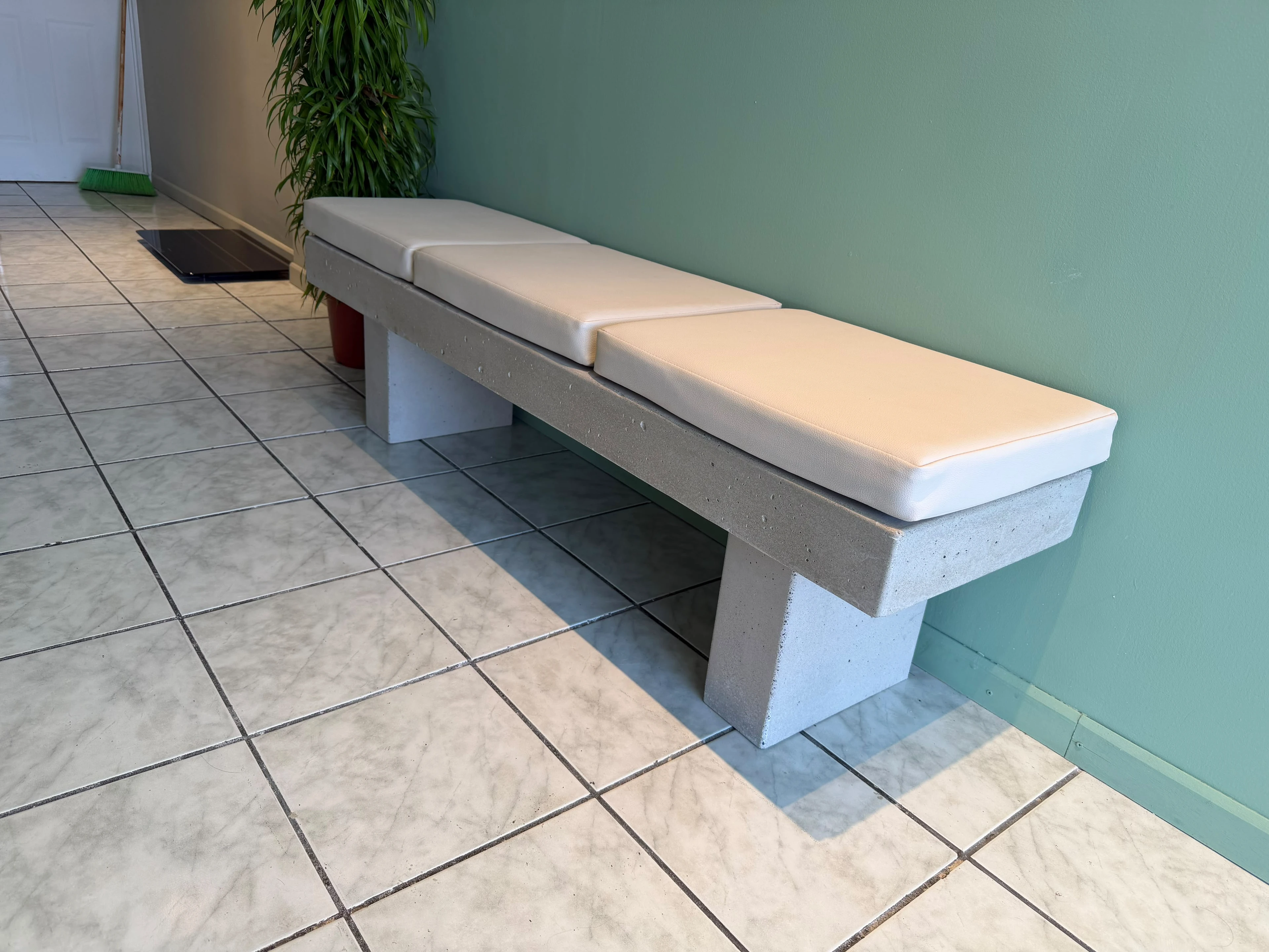 Banca de concreto gris con patas laterales inclinadas y tres cojines blancos, en pasillo con piso de cerámica y pared verde
