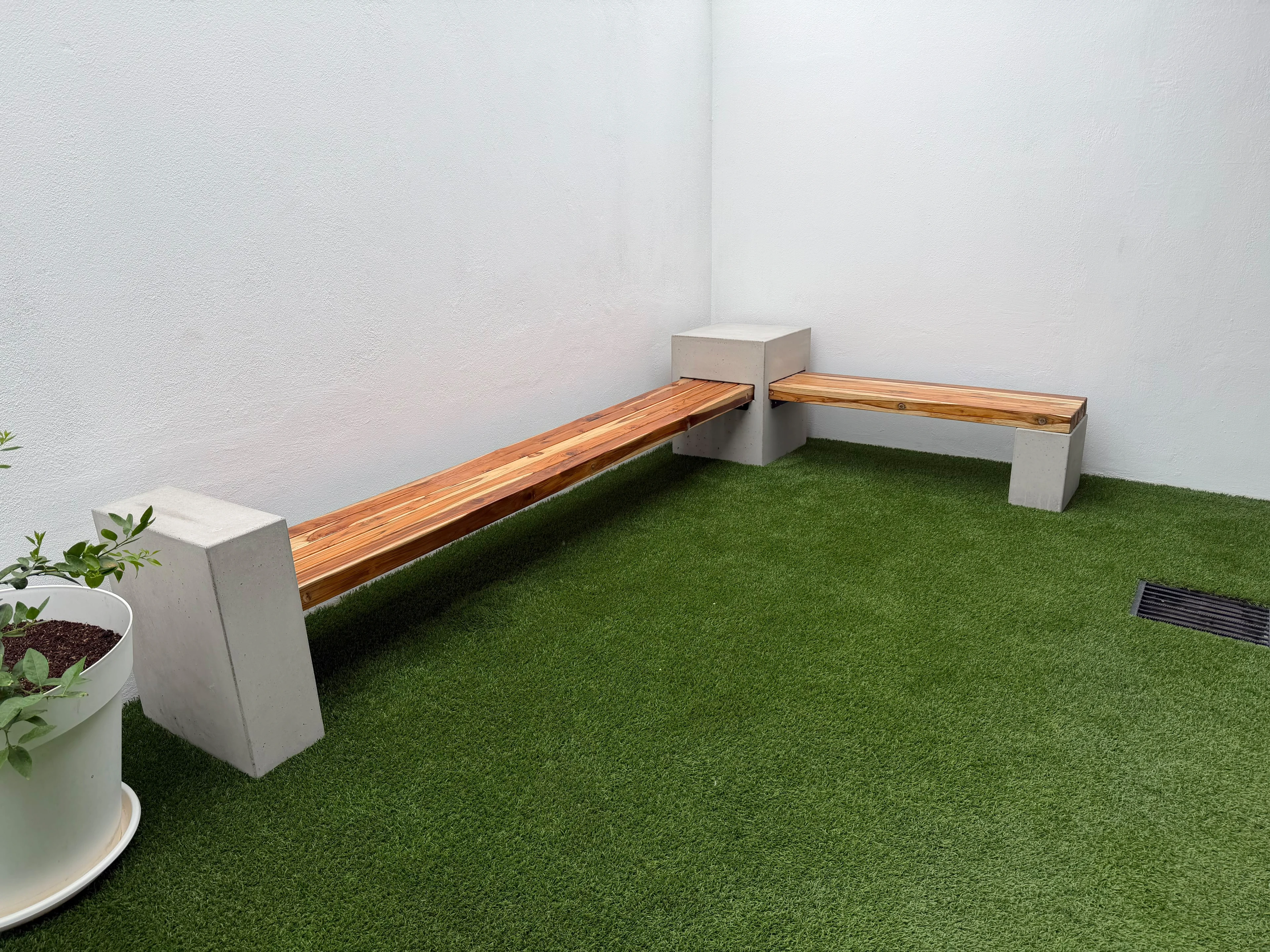 Banca esquinera de concreto con asiento de madera y bloques de concreto como soportes, sobre césped sintético en patio blanco