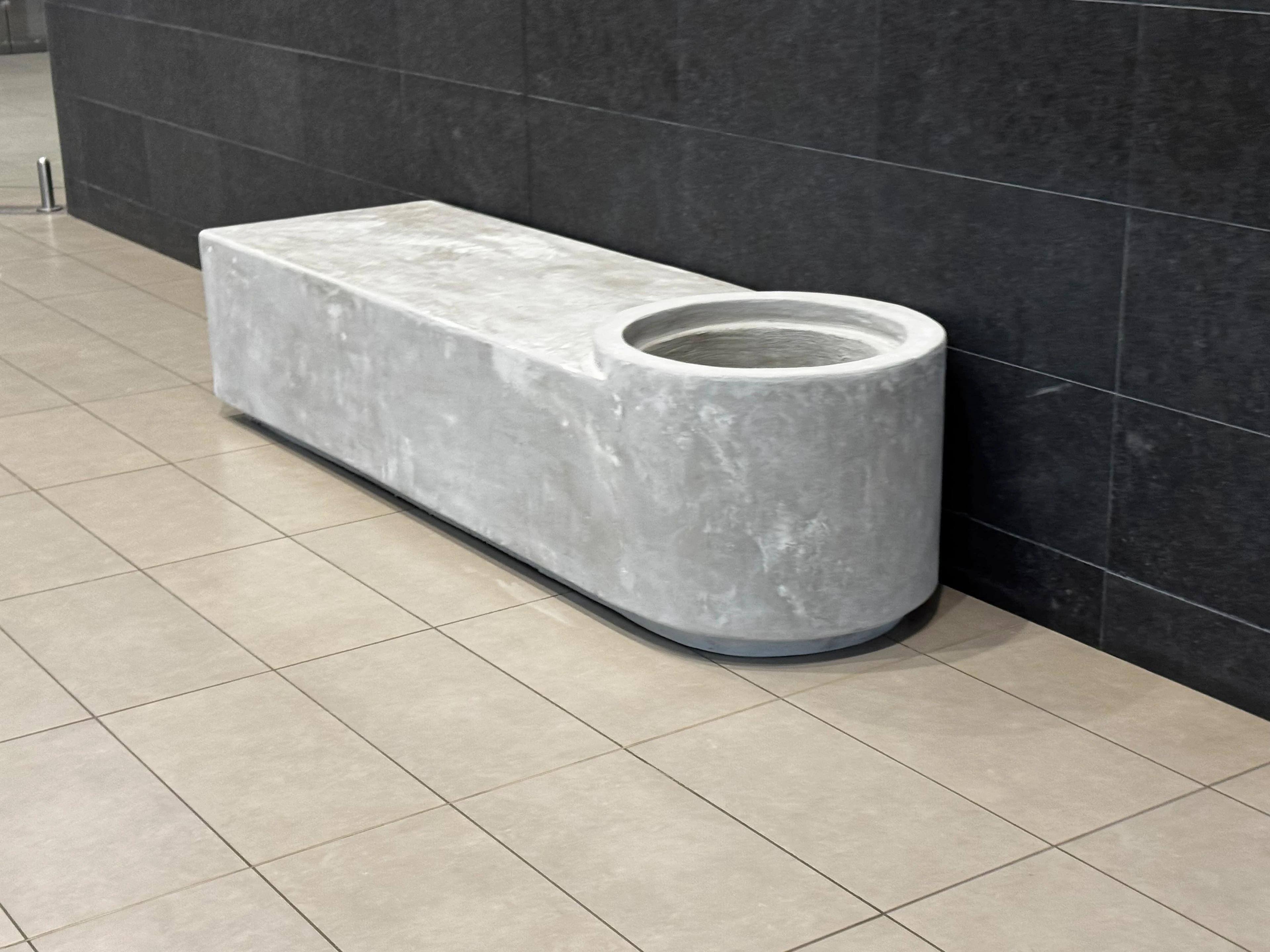 Banca larga de concreto gris con un hueco circular en un extremo, junto a pared de piedra oscura y piso de cerámica