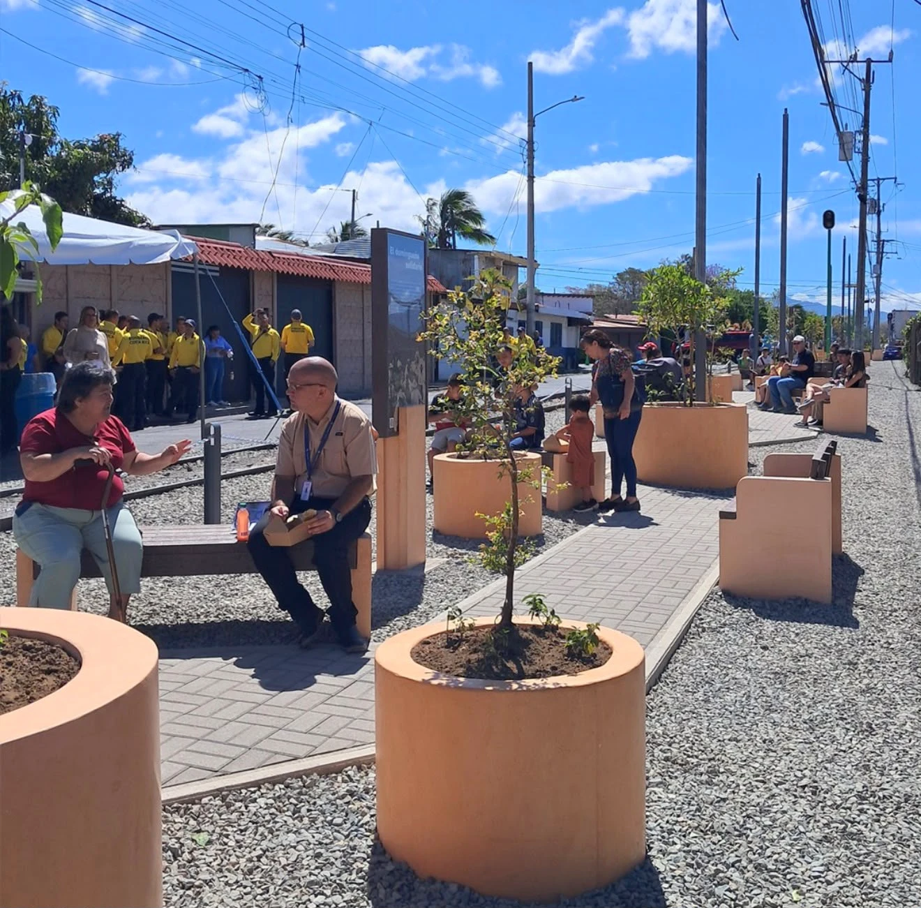 Maceteros cilíndricos de concreto color durazno con árboles jóvenes y bancas del mismo tono, en un paseo peatonal con gente
