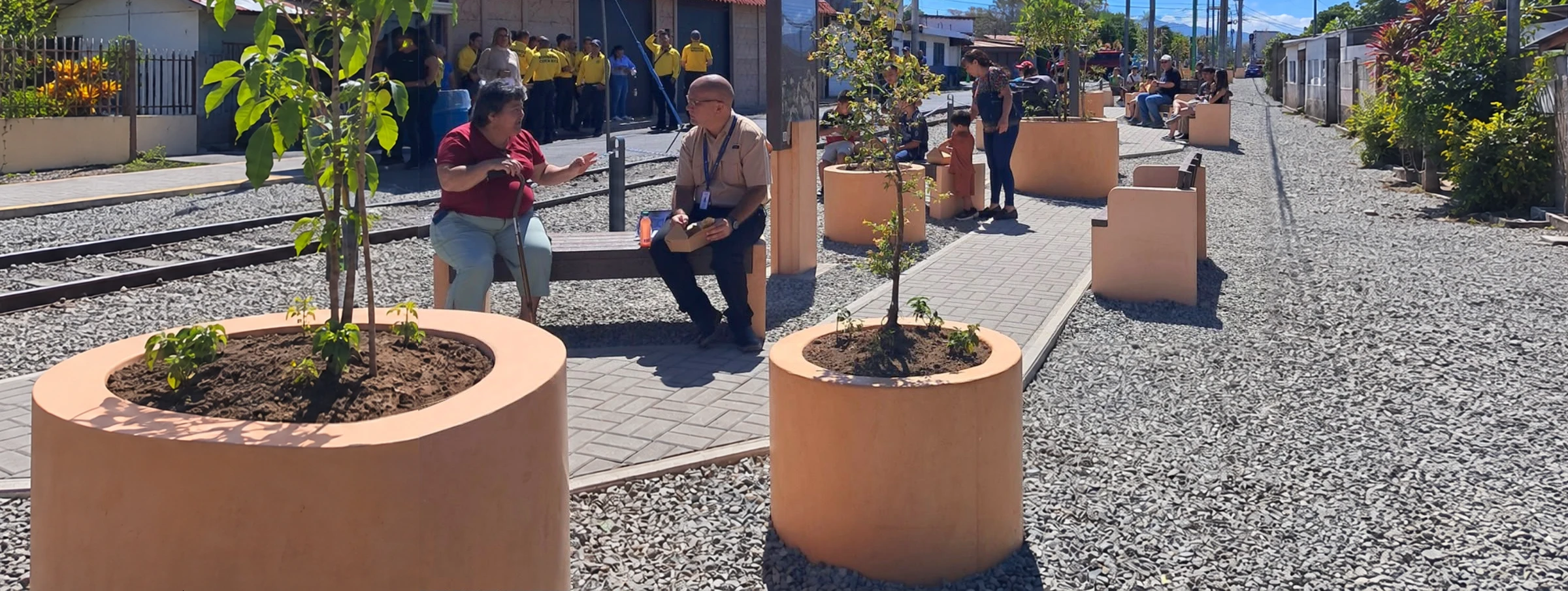 Bancas y jardineras de concreto color terracota en un paseo junto a la vía del tren, con personas disfrutando del espacio