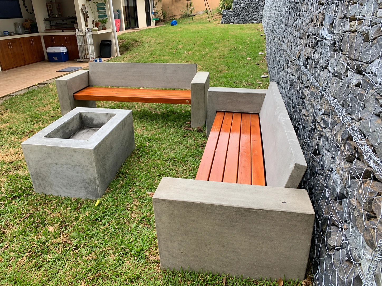 Bancas de concreto gris con asientos de listones de madera y una jardinera rectangular, sobre césped junto a un muro de gaviones