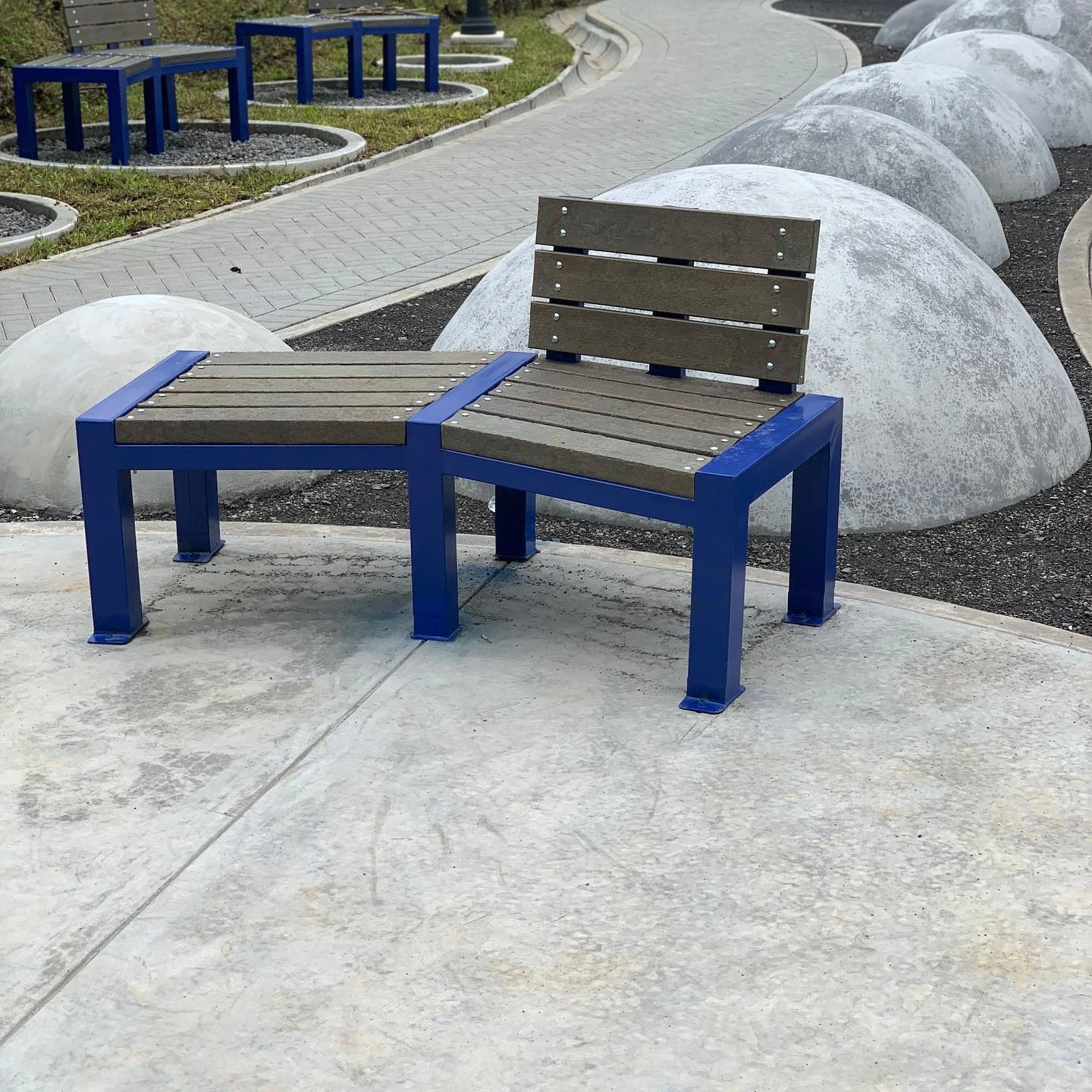 Banca de listones de madera sobre estructura azul junto a un sendero, con esferas de concreto al fondo