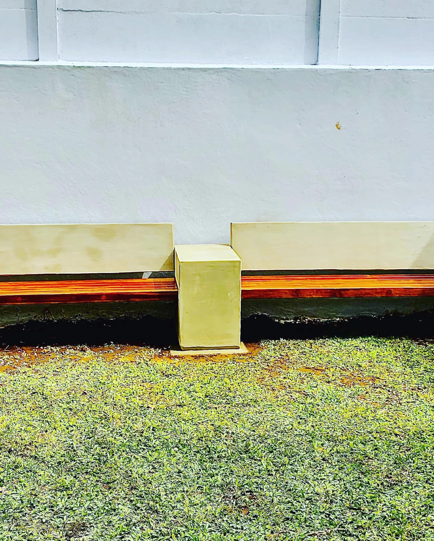 Banca larga de concreto claro con respaldo y listones de madera anaranjada, contra una pared blanca sobre césped. Vista adicional 1.