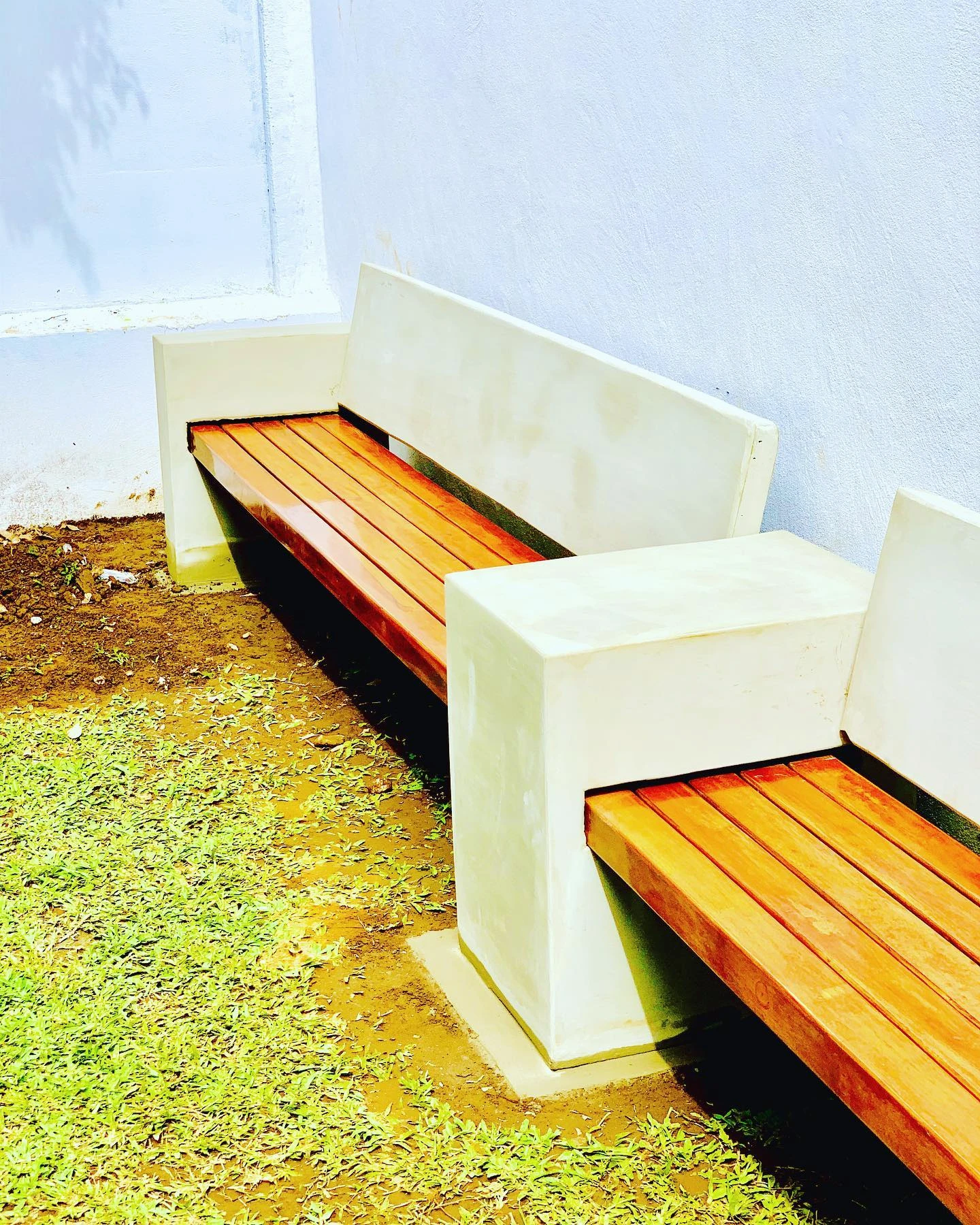 Banca larga de concreto claro con respaldo y listones de madera anaranjada, contra una pared blanca sobre césped. Vista adicional 2.