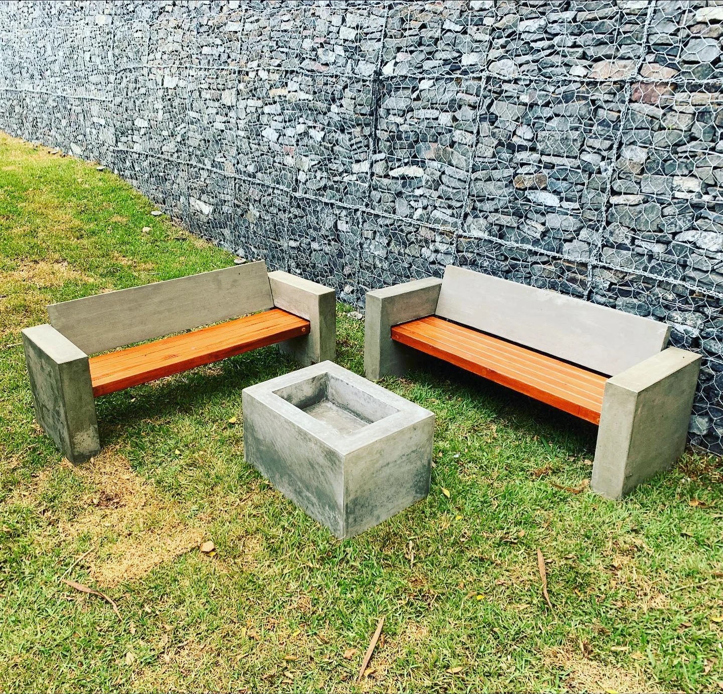Banca de concreto con listones de madera anaranjada junto a una jardinera cuadrada, contra un muro de gaviones