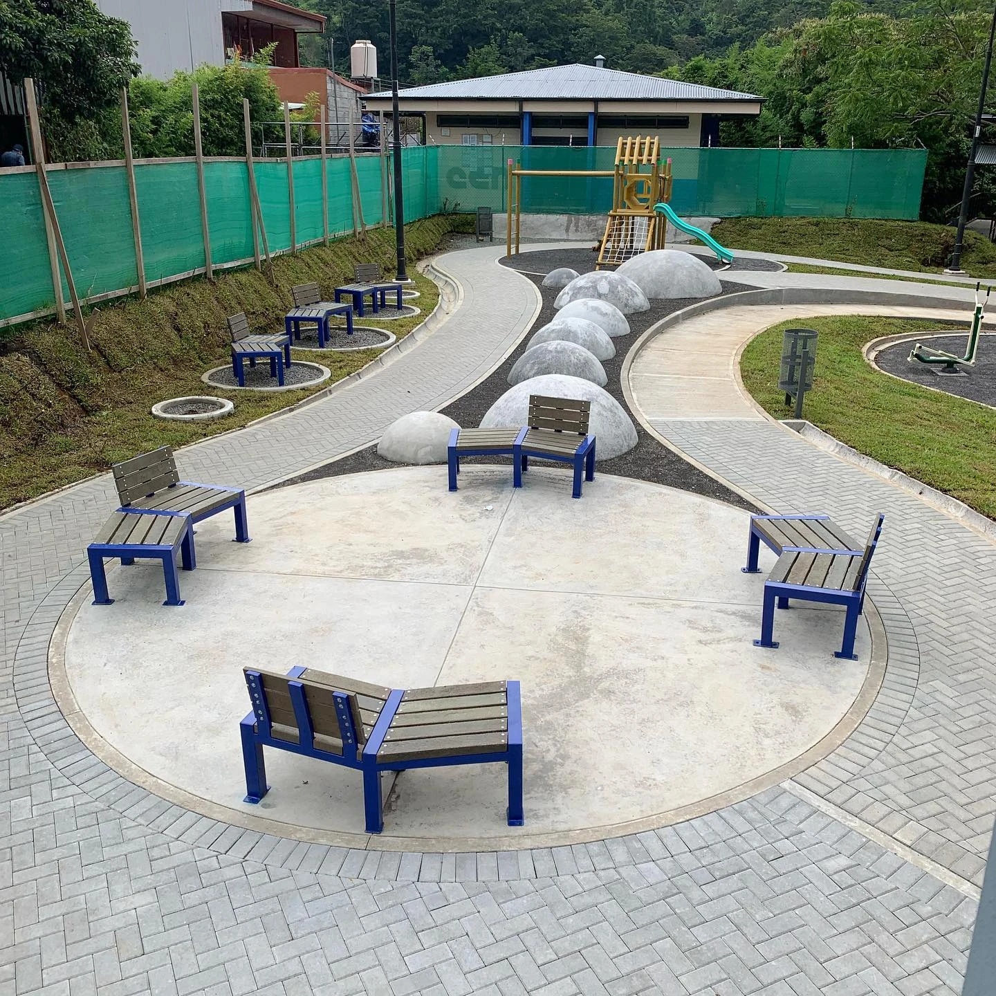 Bancas de concreto con listones de madera y estructura azul, distribuidas en un parque con piso de adoquín