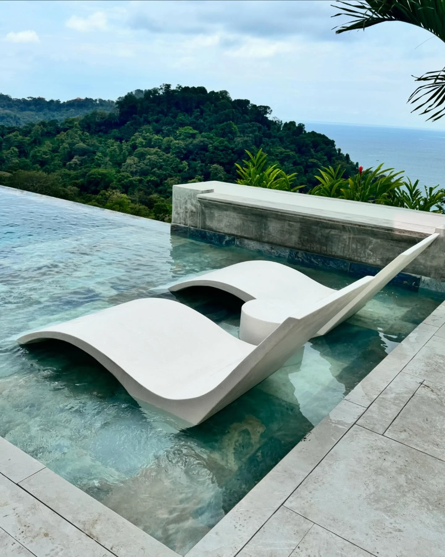 Dos camastros curvos de concreto blanco dentro de una piscina poco profunda, con vista al mar y vegetación