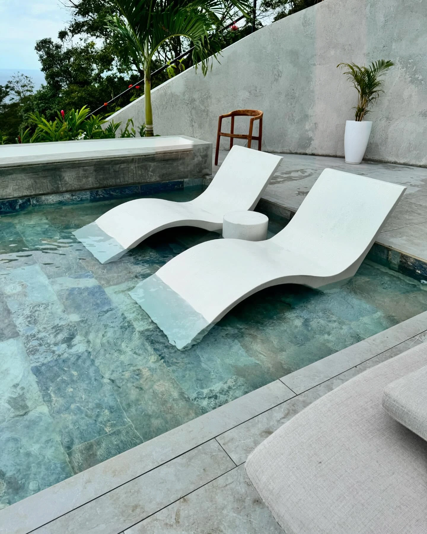 Dos camastros curvos de concreto blanco dentro de una piscina poco profunda, con vista al mar y vegetación. Vista adicional 1.