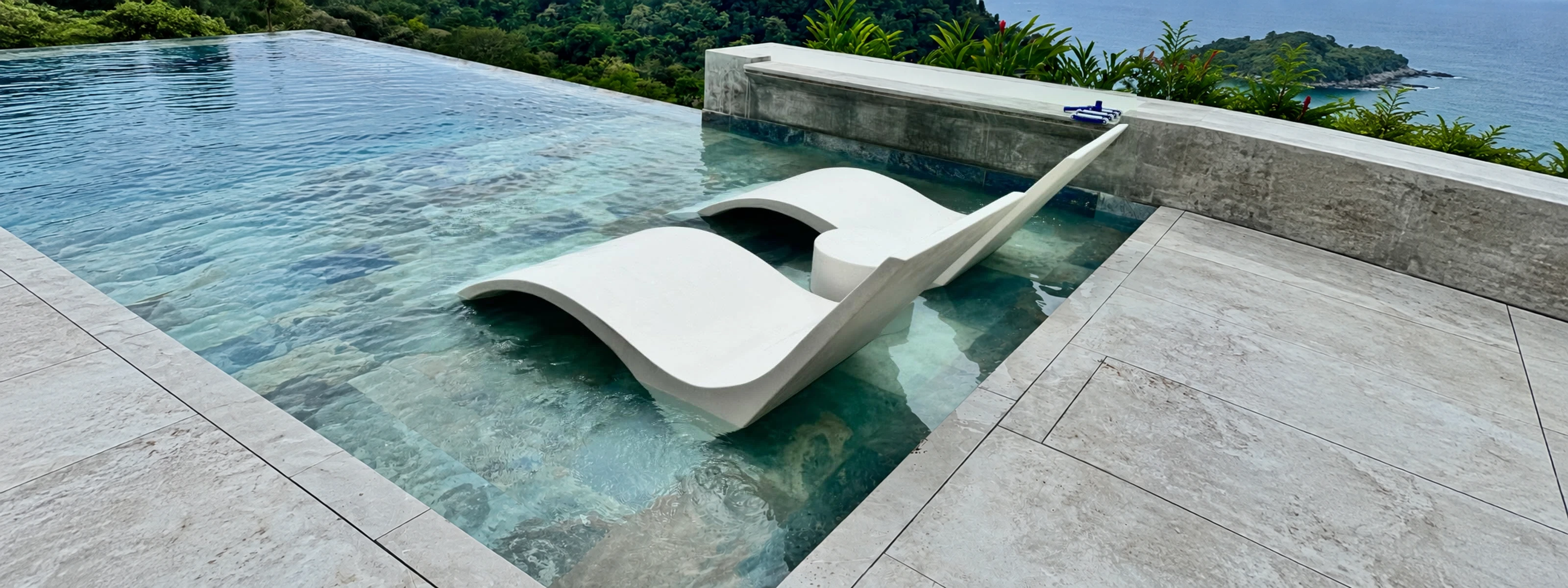 Dos camastros curvos de concreto blanco dentro de una piscina poco profunda, con vista al mar
