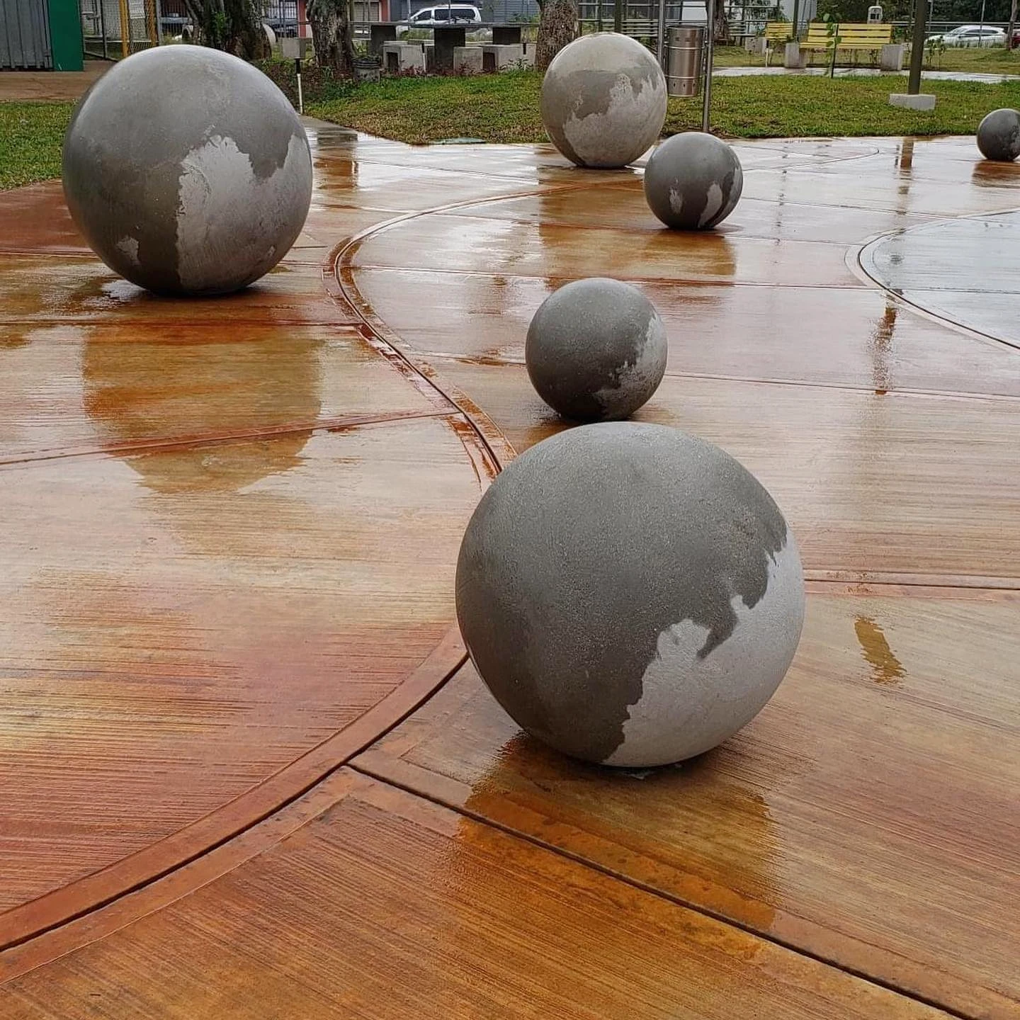 Cuatro esferas de concreto gris con relieve de continentes, distribuidas sobre el piso rojizo húmedo de una plaza