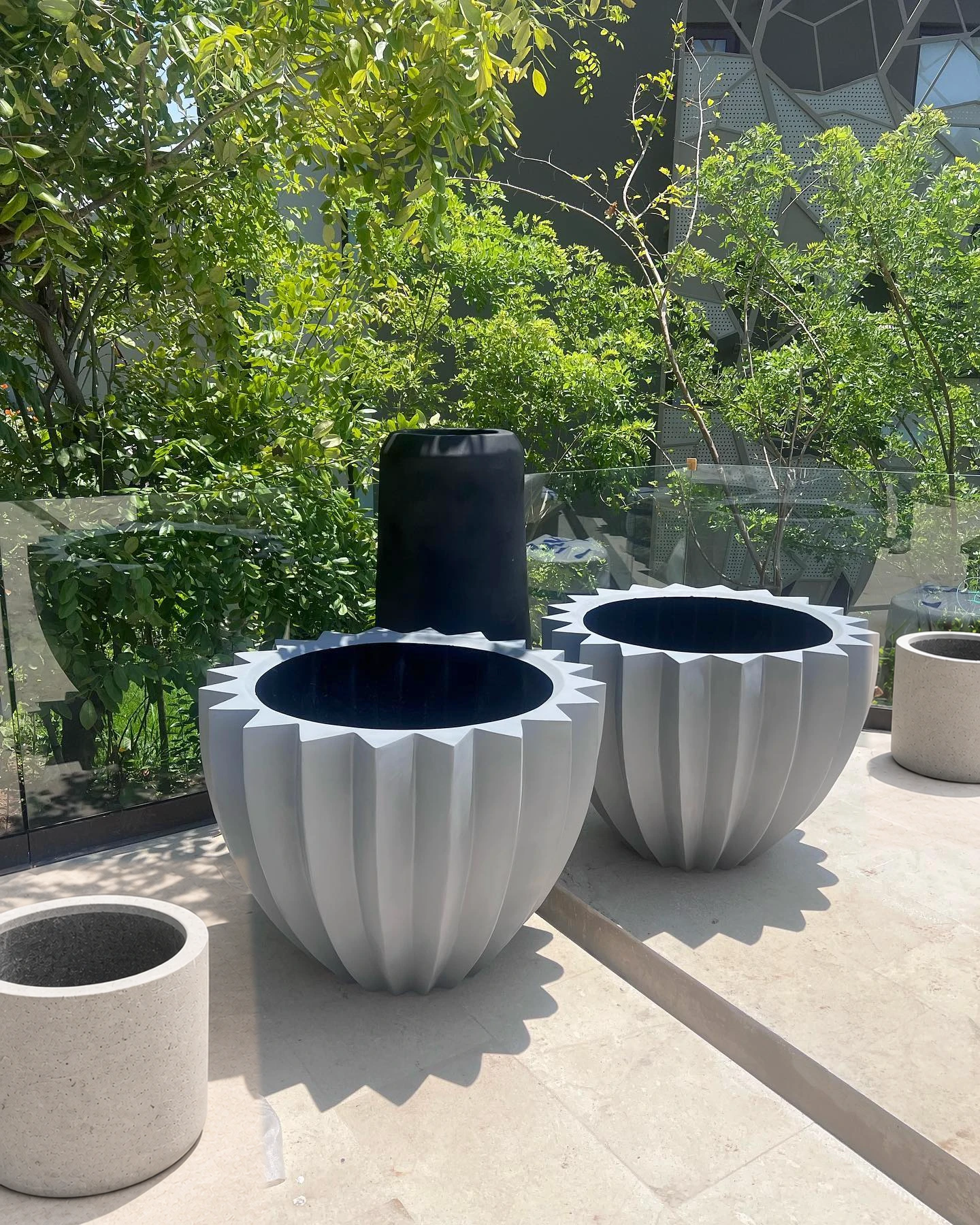 Dos maceteros de concreto claro con estriado vertical y un macetero negro alto, en una terraza