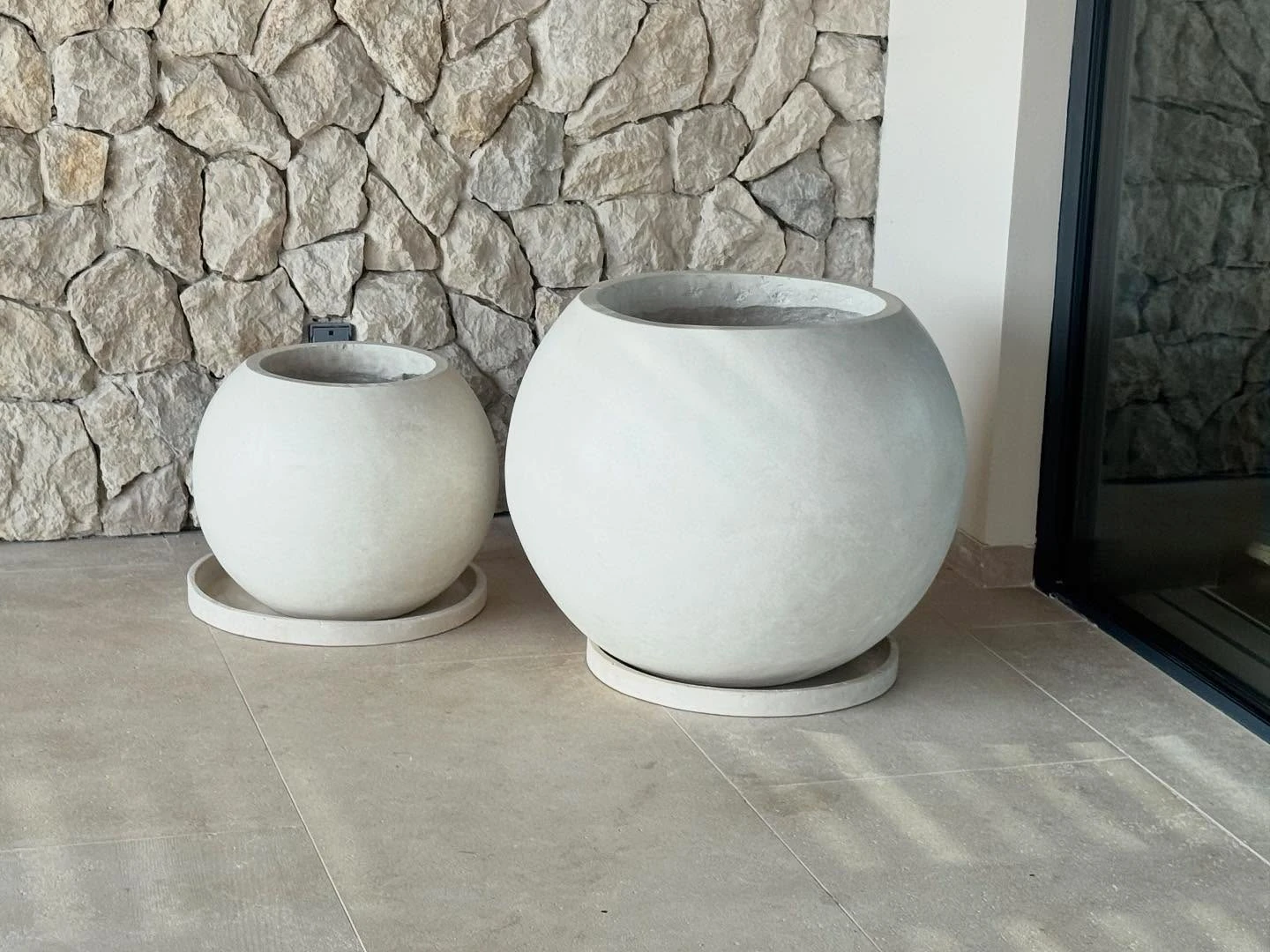 Dos maceteros esféricos de concreto claro sobre platos, contra un muro de piedra apilada