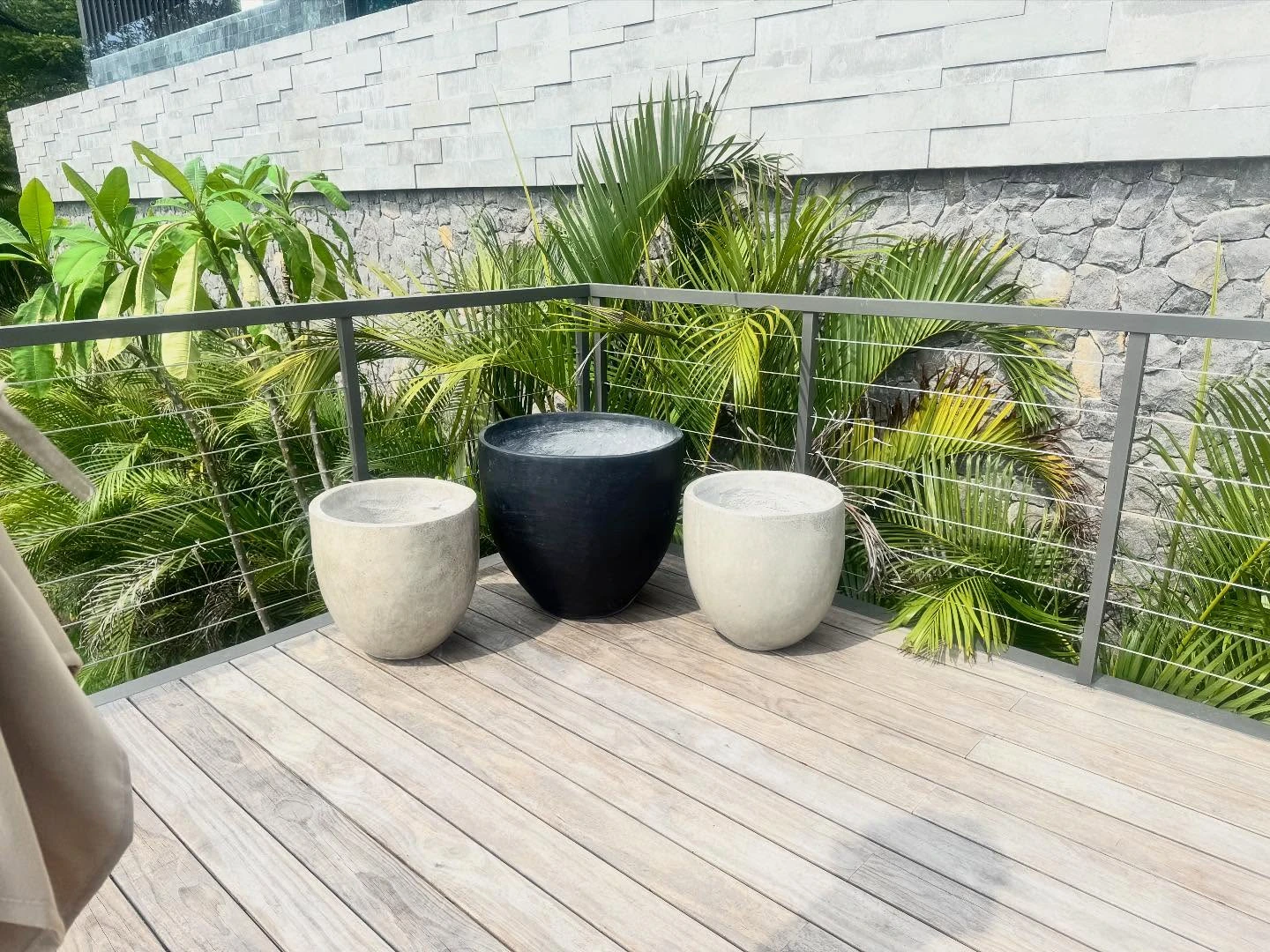 Tres maceteros de concreto, dos claros y uno negro, sobre un deck de teca rodeado de palmas