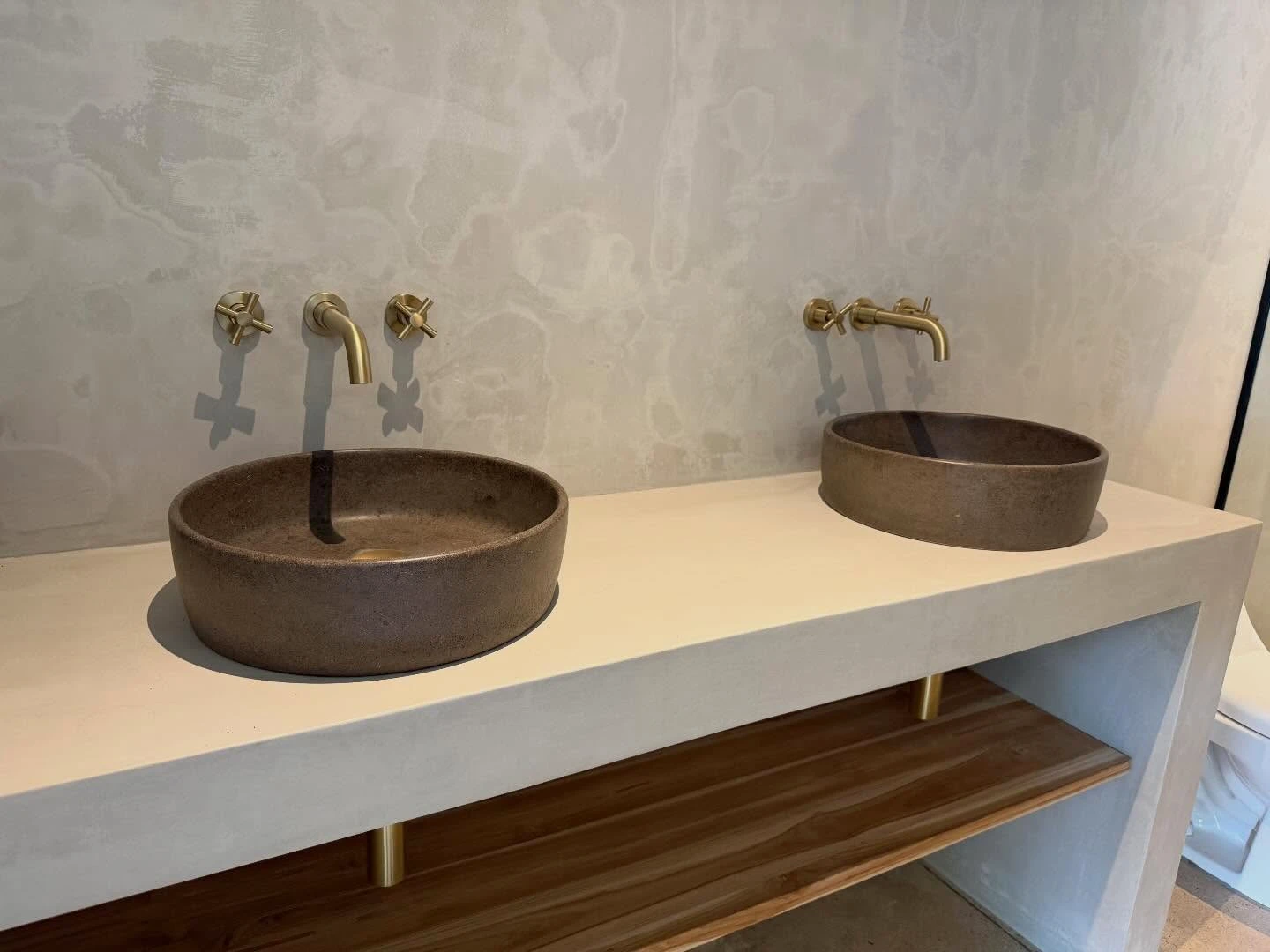 Dos lavatorios redondos de concreto color bronce sobre un sobre blanco con repisa de madera y grifería dorada de pared