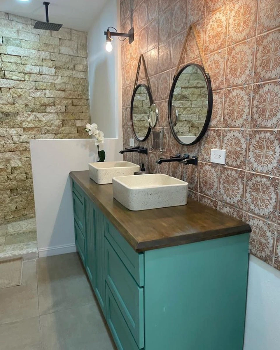 Dos lavatorios cuadrados de concreto claro sobre un mueble turquesa con sobre de madera, contra una pared de azulejo terracota estampado