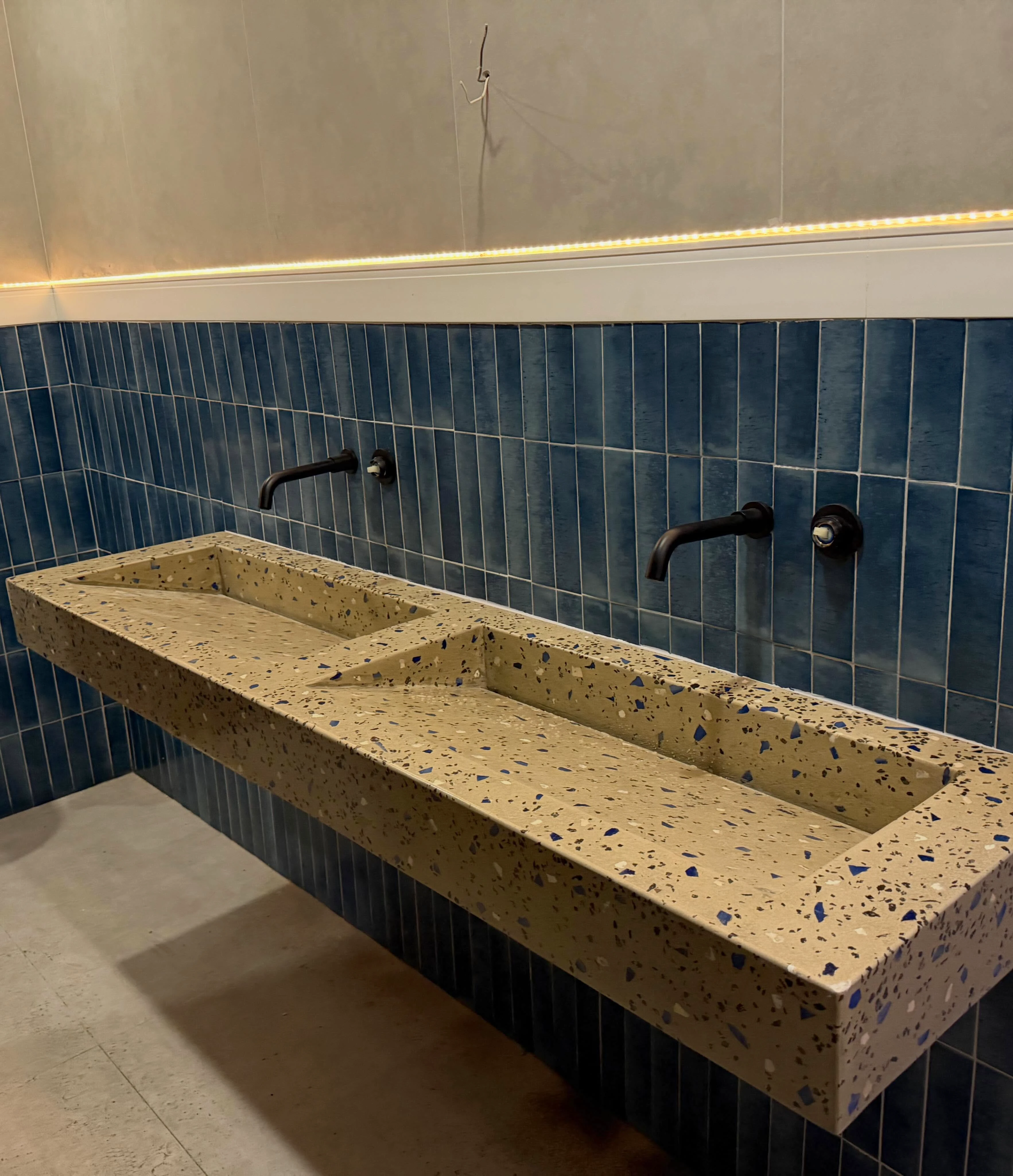 Lavatorio lineal de terrazo con dos pozas rectangulares, sobre pared de azulejo azul oscuro