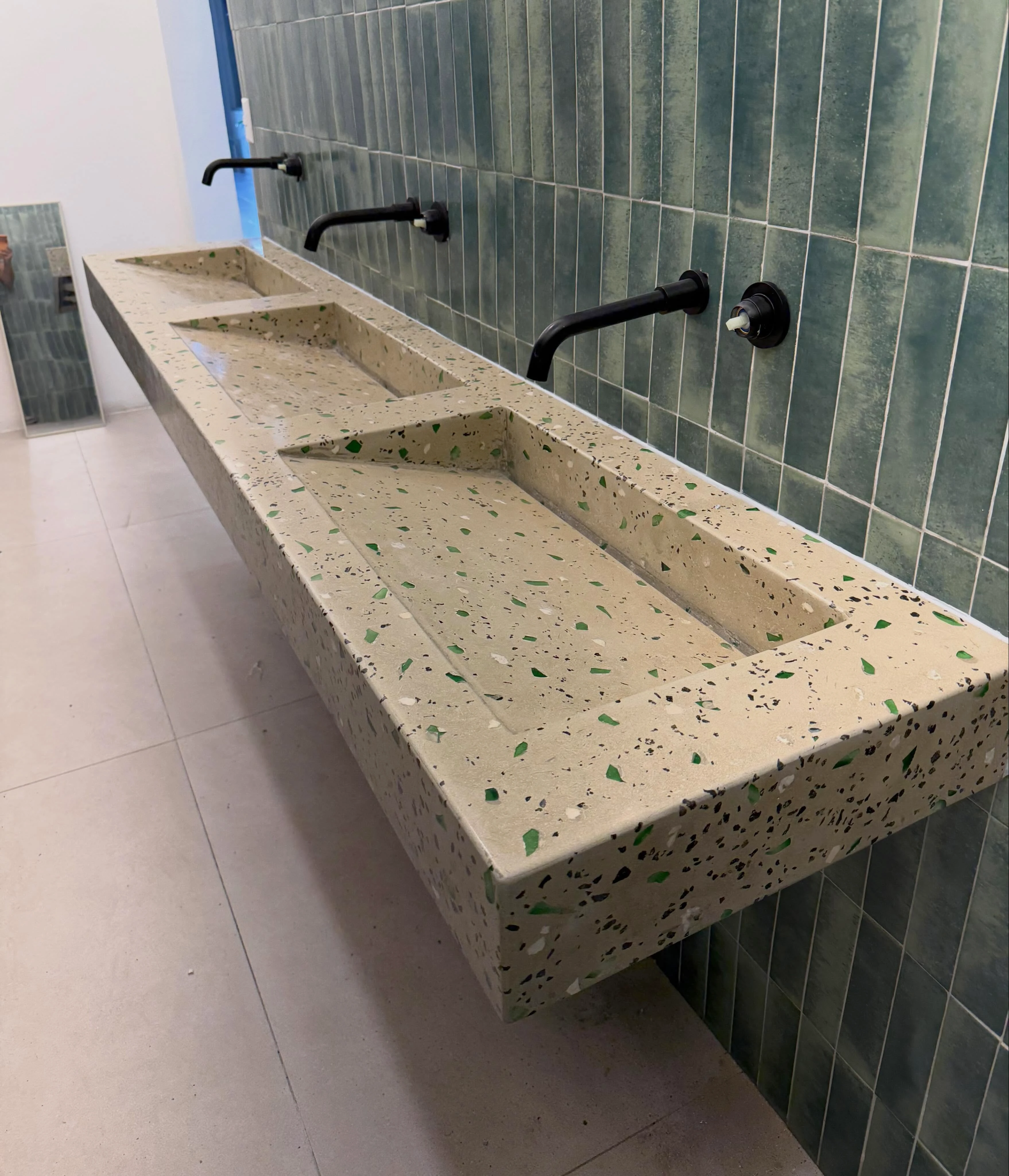 Lavatorio corrido de terrazo claro con agregado de vidrio verde, montado sobre pared de azulejo verde con grifería negra de pared