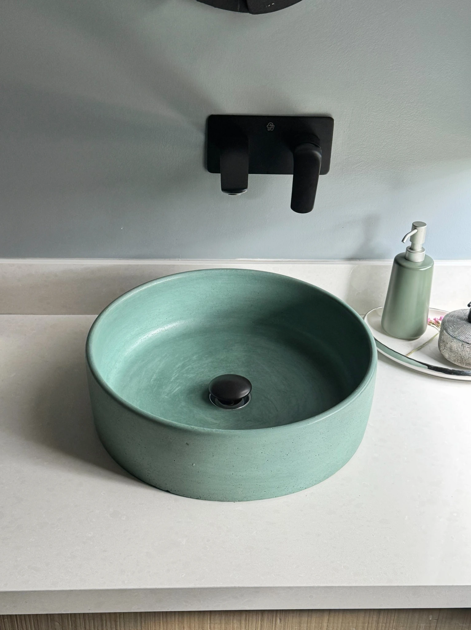 Lavatorio redondo de concreto verde menta sobre un sobre claro, con grifería negra de pared y muro gris
