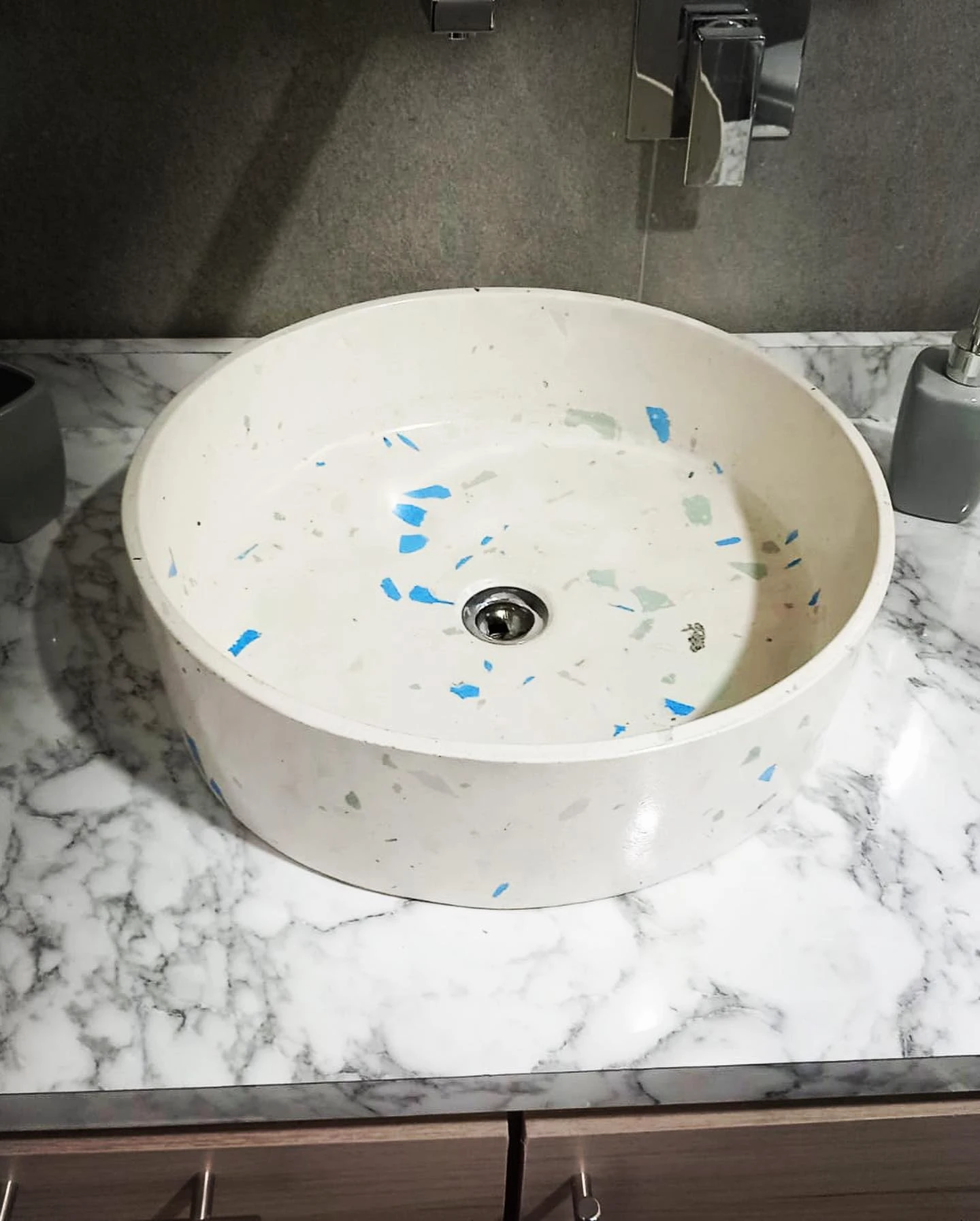 Lavatorio redondo de terrazo blanco con fragmentos de vidrio azul, apoyado sobre un sobre de mármol blanco