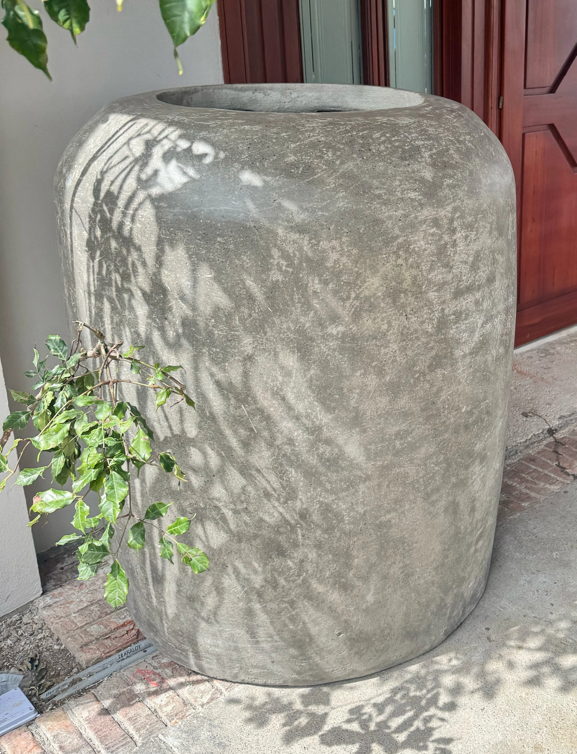 Macetero cilíndrico alto de concreto gris con hombro redondeado, junto a la puerta de madera de una entrada