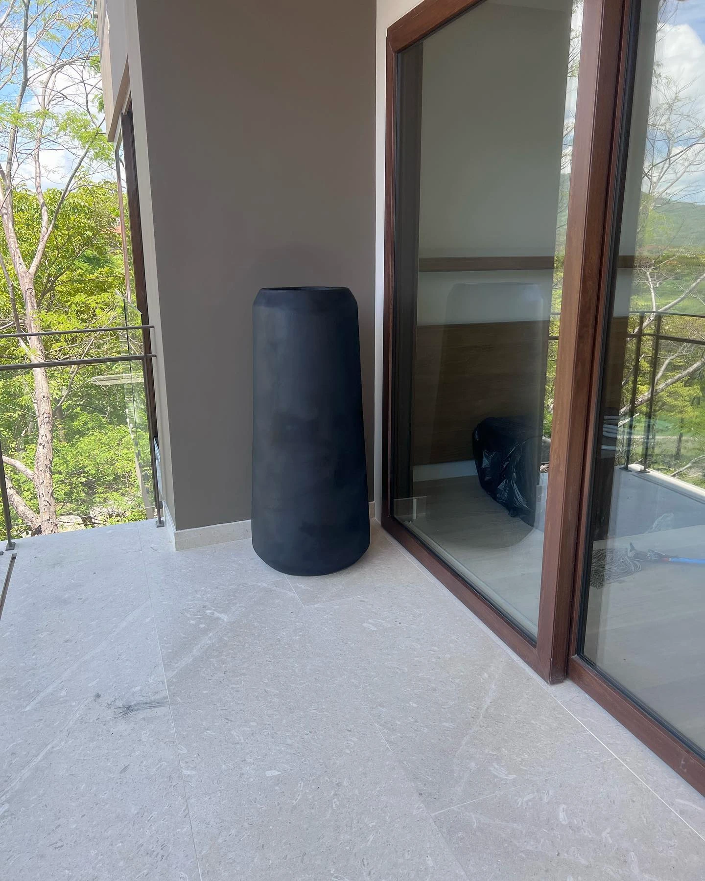 Macetero cónico alto de concreto negro junto a un ventanal, con vegetación al fondo