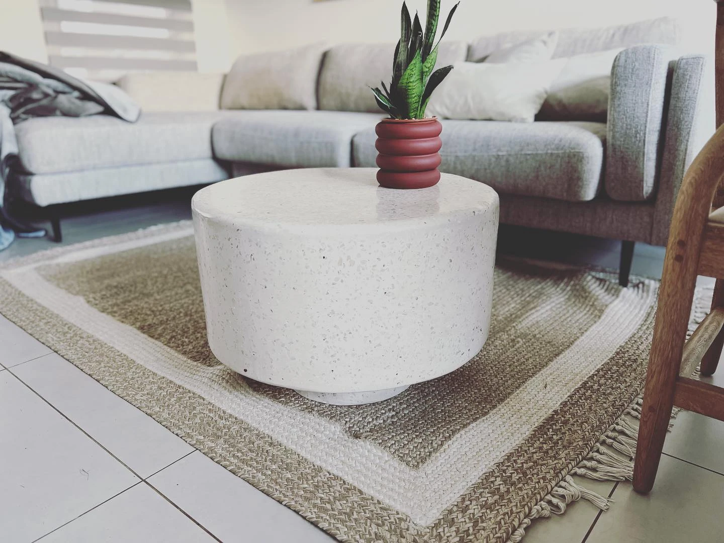 Mesa de centro cilíndrica de terrazo claro con agregado oscuro, sobre una alfombra junto a un sofá blanco