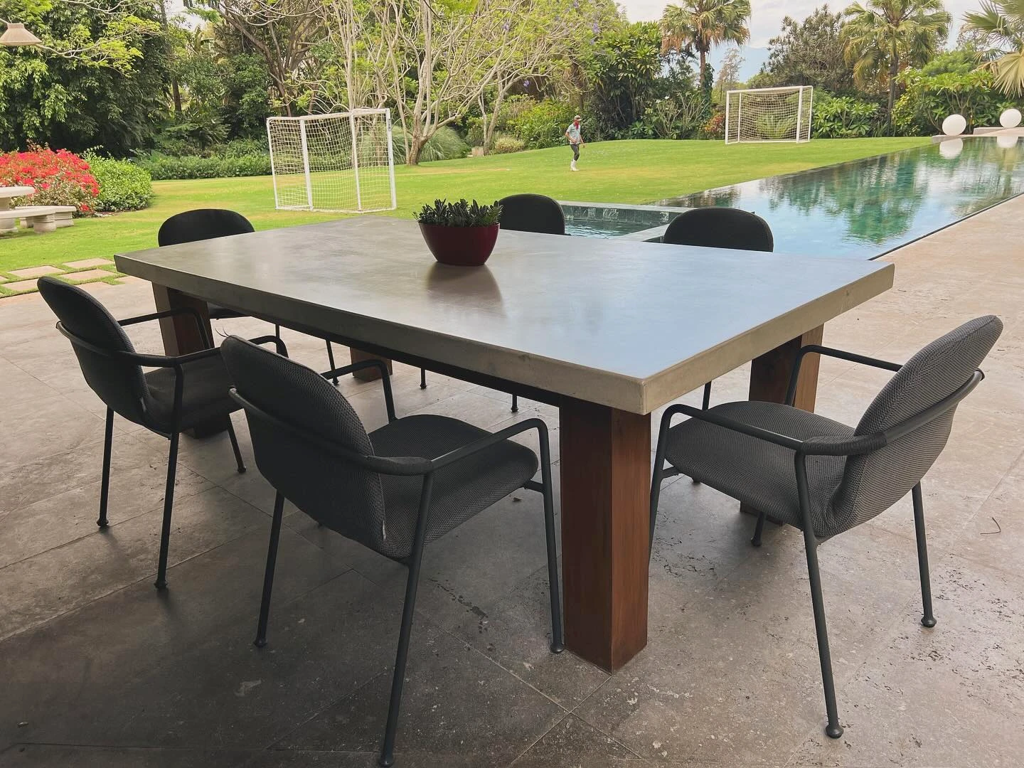 Mesa de comedor larga con sobre de concreto gris y patas de madera de teca, en una terraza frente a un jardín