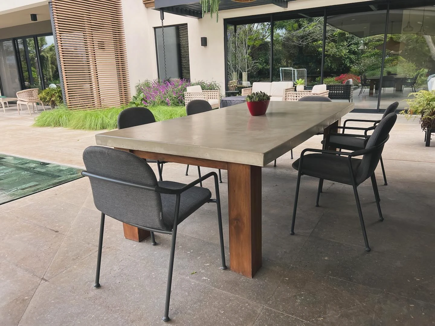 Mesa de comedor larga con sobre de concreto gris y patas de madera de teca, en una terraza frente a un jardín. Vista adicional 1.