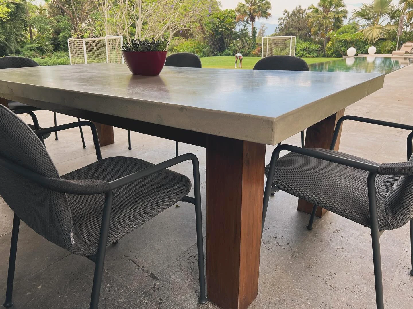 Mesa de comedor larga con sobre de concreto gris y patas de madera de teca, en una terraza frente a un jardín. Vista adicional 2.