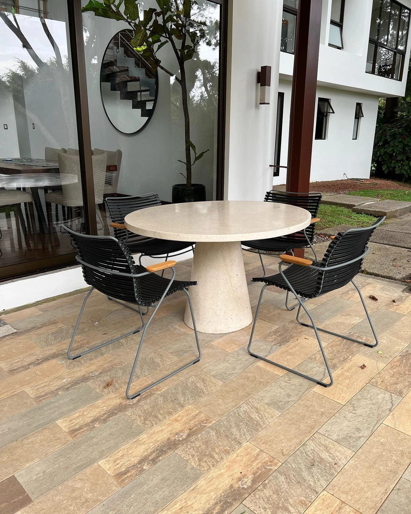 Mesa redonda de concreto claro sobre base cónica, con sillas de alambre negro en una terraza de madera