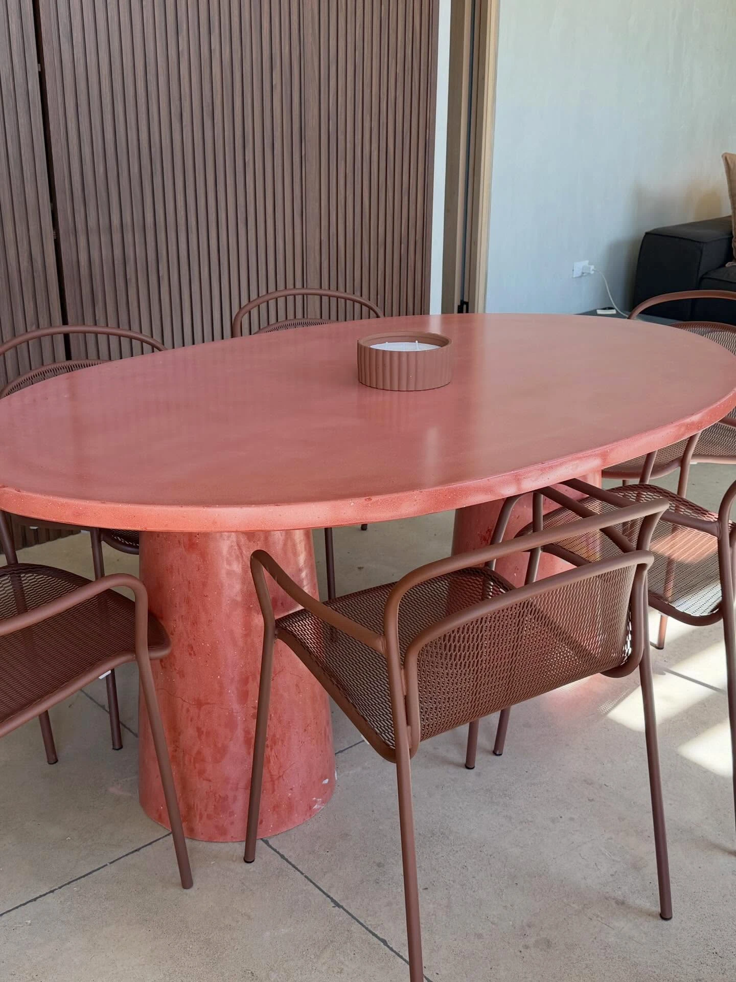 Mesa de comedor ovalada de concreto terracota sobre base cónica, con seis sillas de metal en el mismo tono