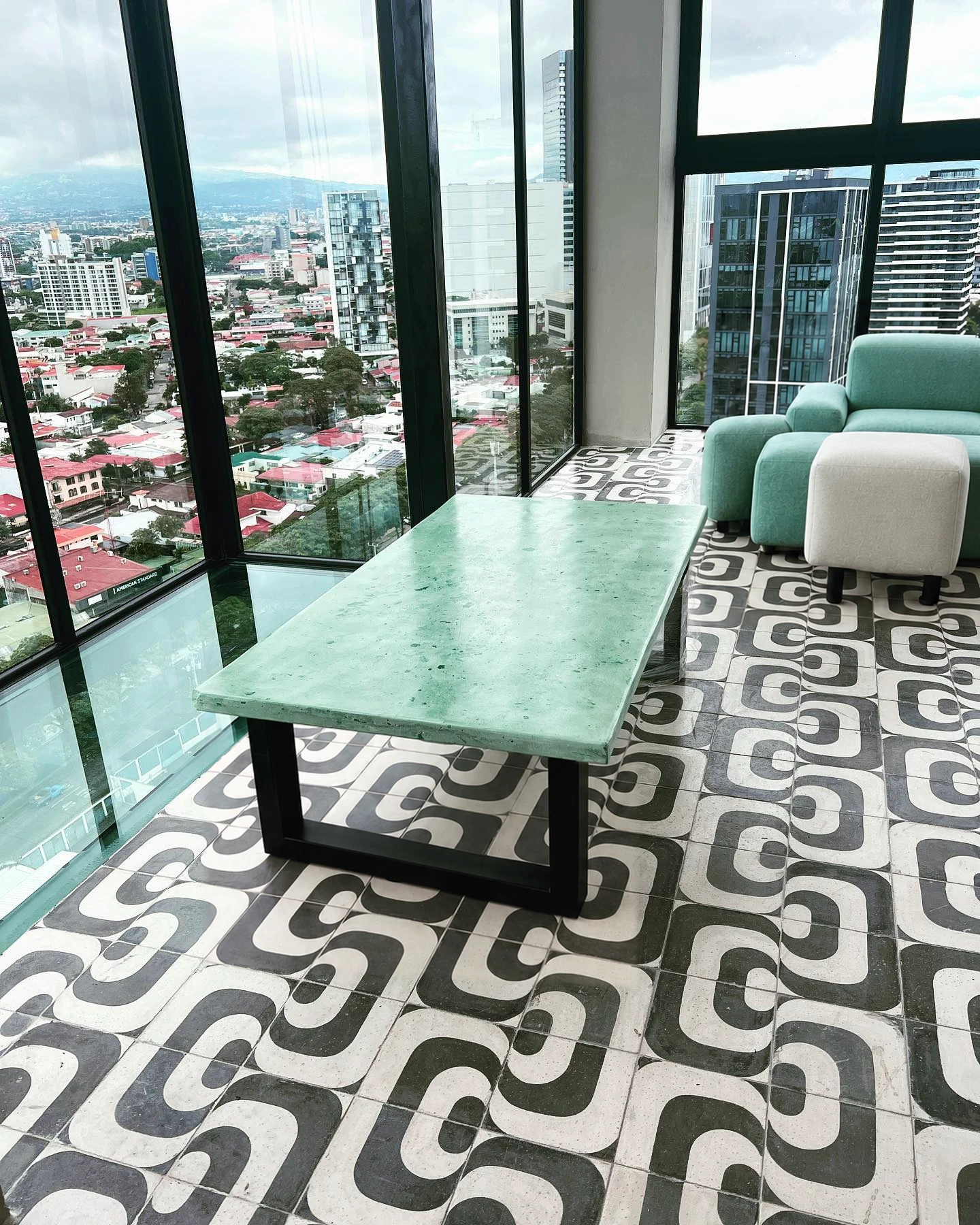 Mesa rectangular de concreto verde marmoleado sobre un piso de mosaico gráfico en blanco y negro, con vista a la ciudad