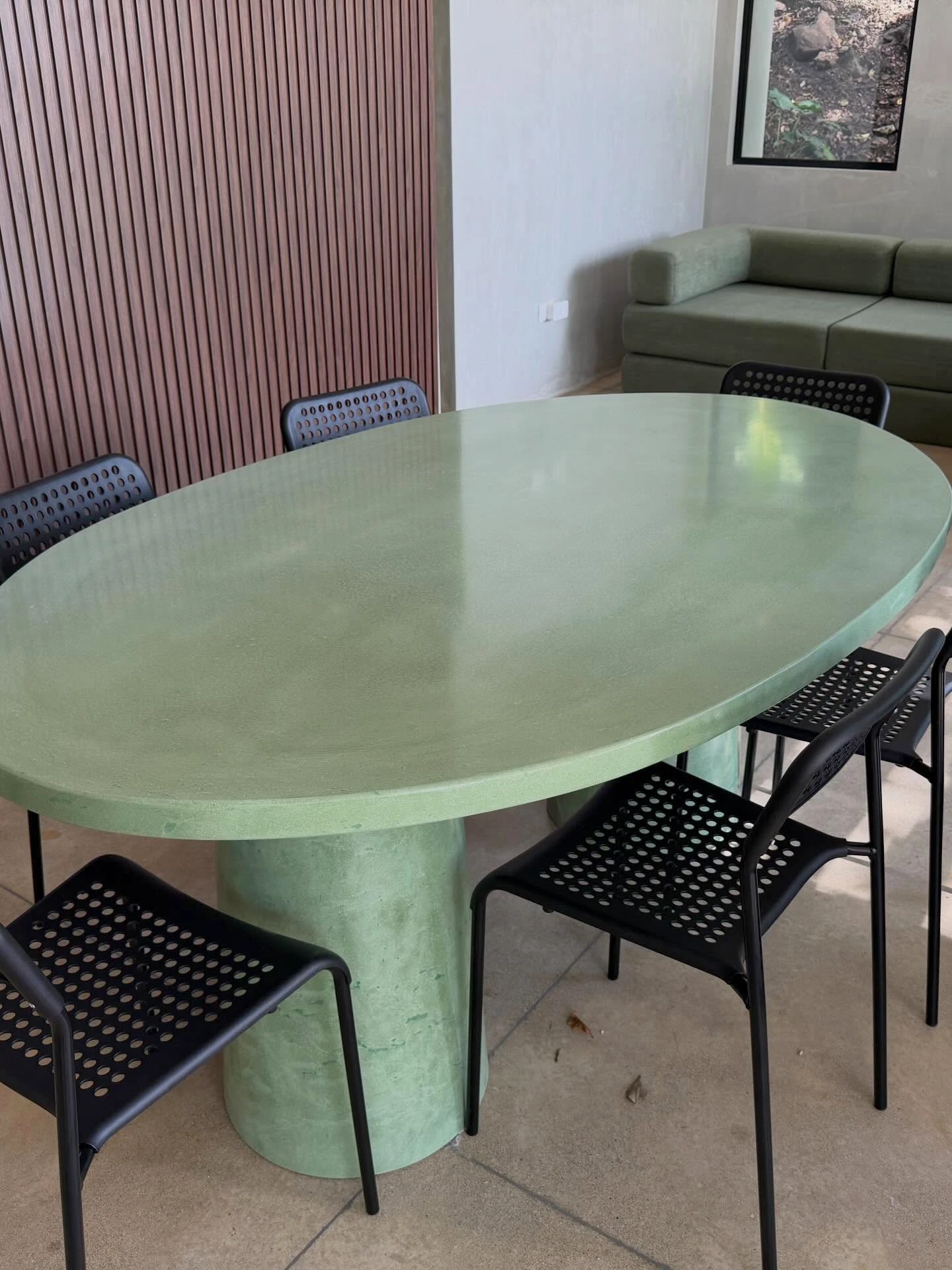Mesa de comedor ovalada de concreto verde salvia con sillas negras, contra una pared de listones de madera