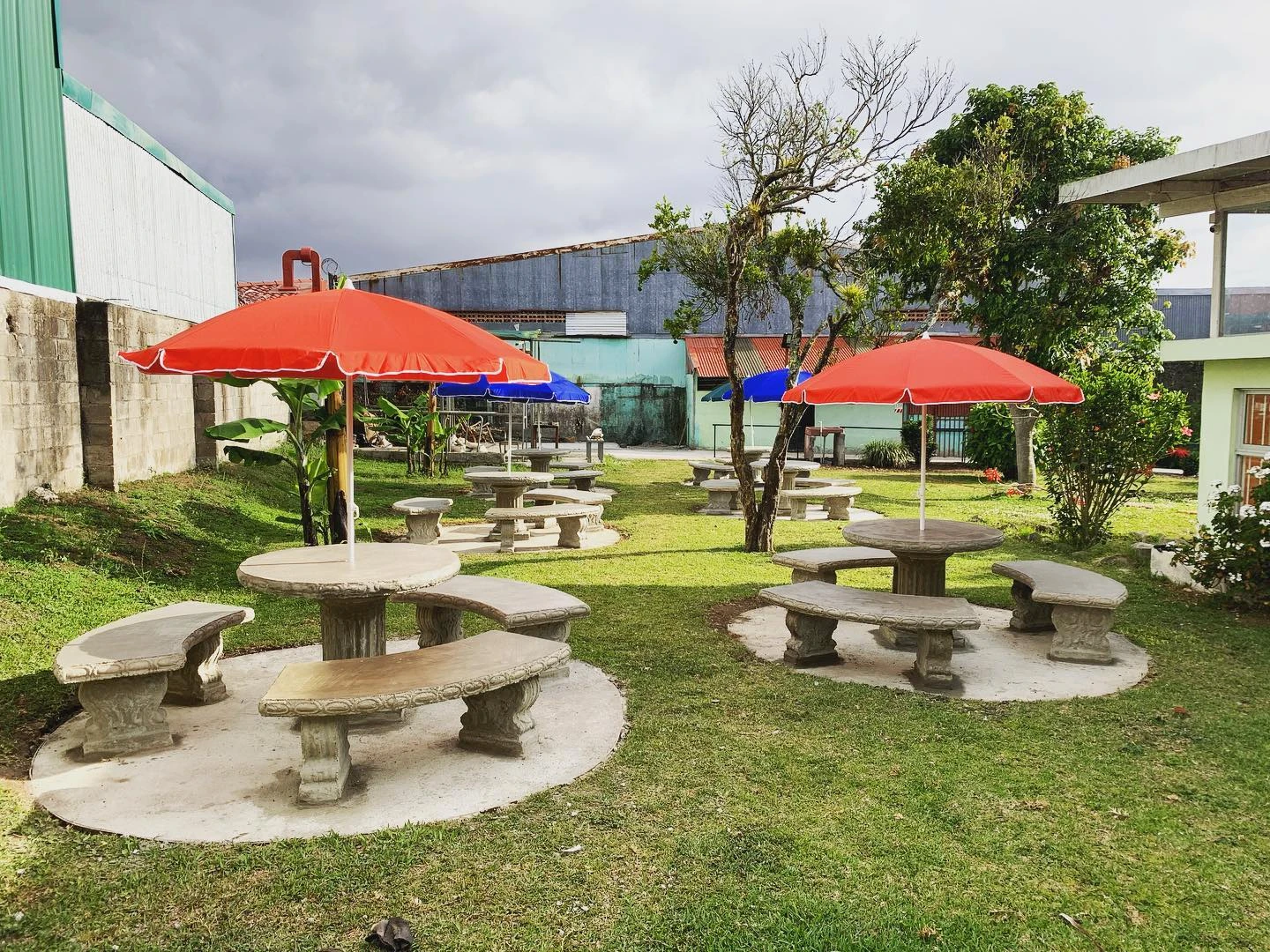 Conjuntos de mesa redonda y bancas de concreto bajo sombrillas rojas, en un parque con césped