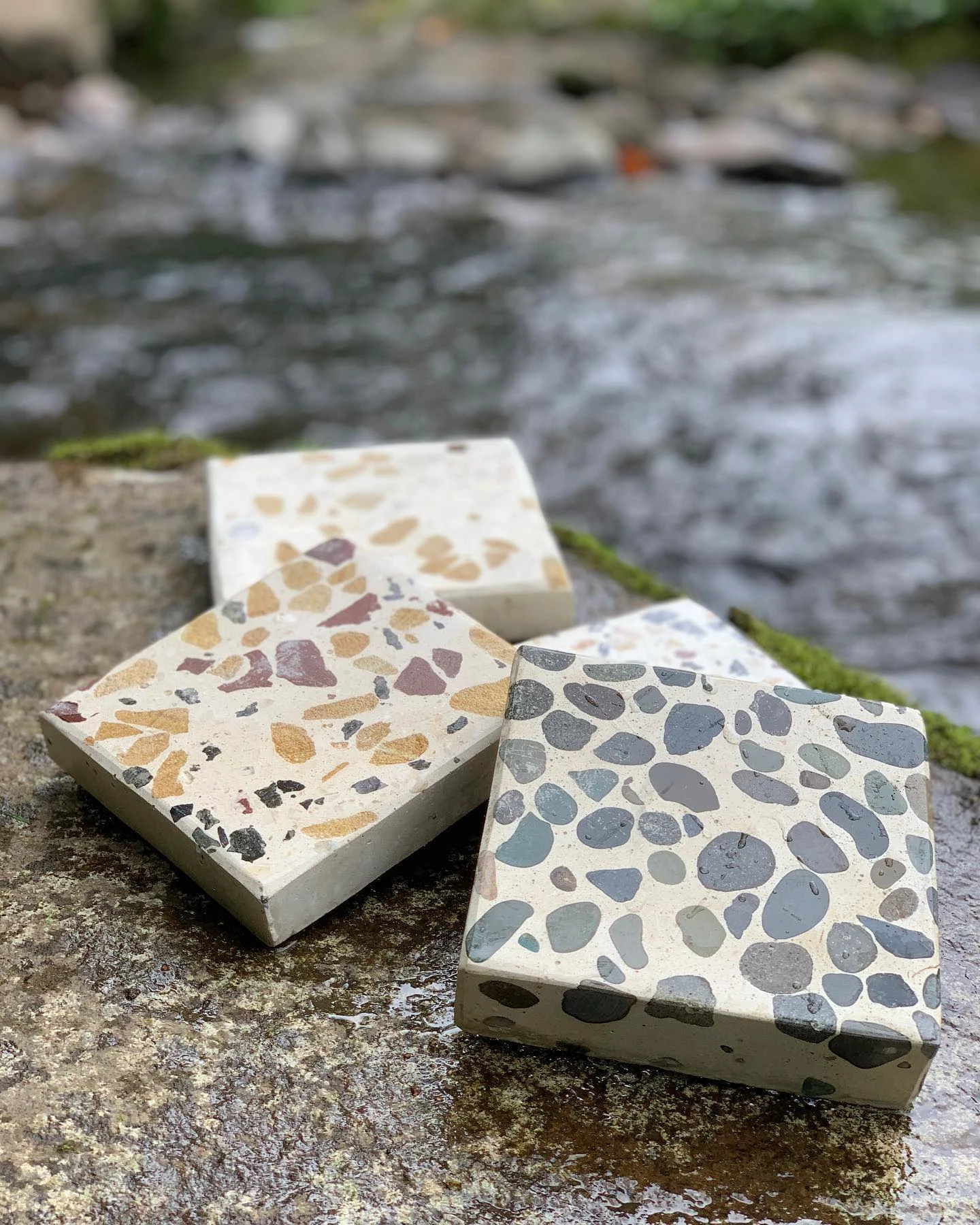 Tres muestras cuadradas de terrazo con agregados de distinto color y tamaño, apoyadas sobre una piedra junto a un río