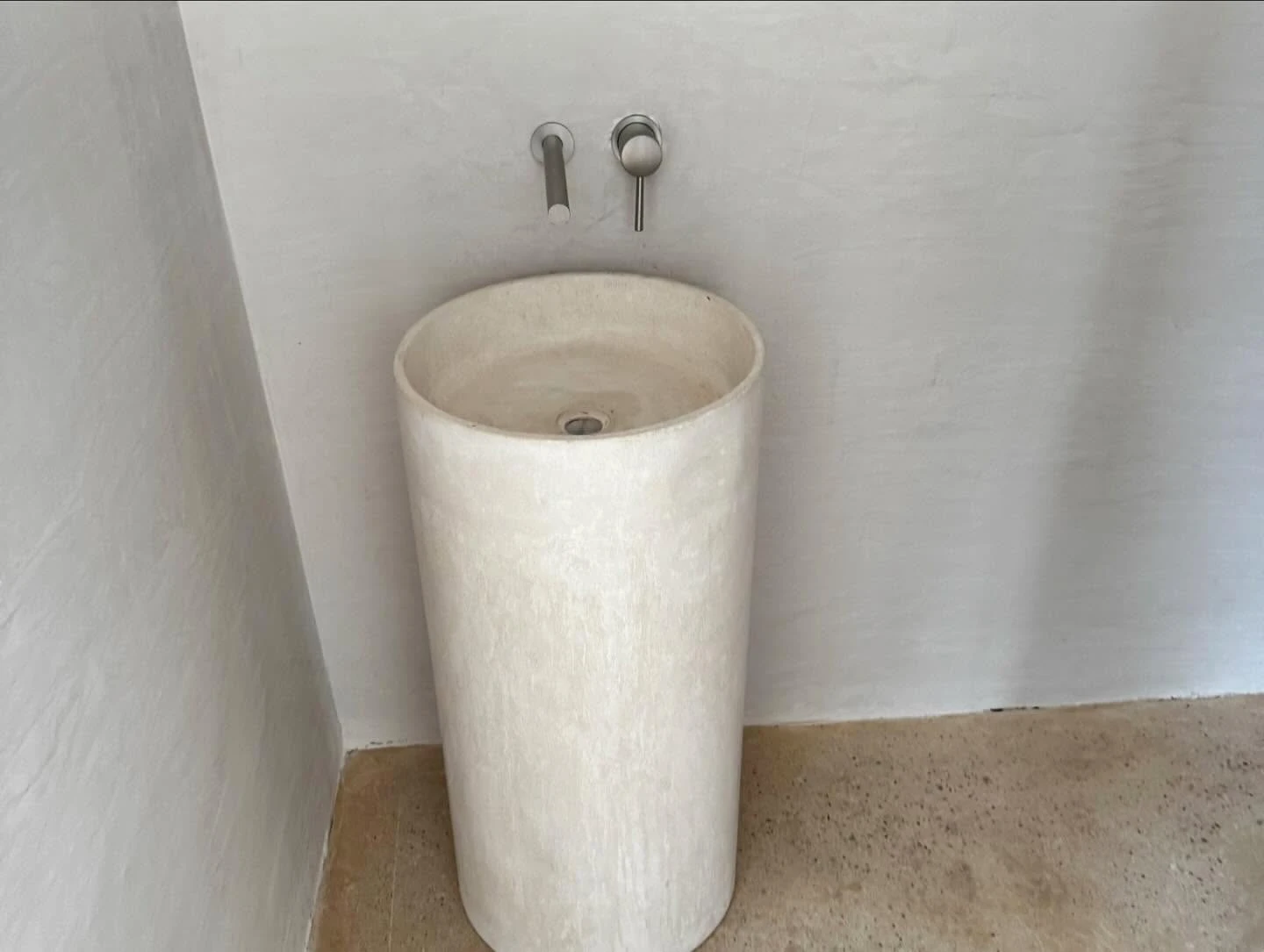 Lavatorio de pedestal cilíndrico en concreto claro, con grifería de pared en acero