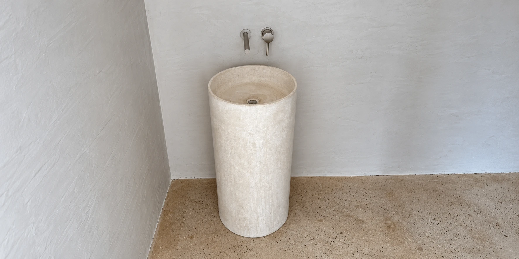 Lavatorio de pedestal cilíndrico de concreto claro con grifería de pared, en un baño blanco