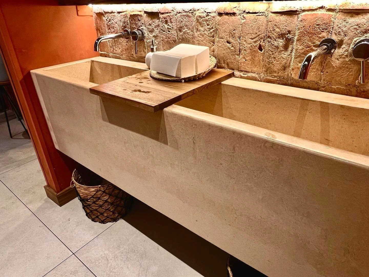 Sobre de concreto claro con poza corrida y bandeja de madera atravesada, contra un muro de piedra