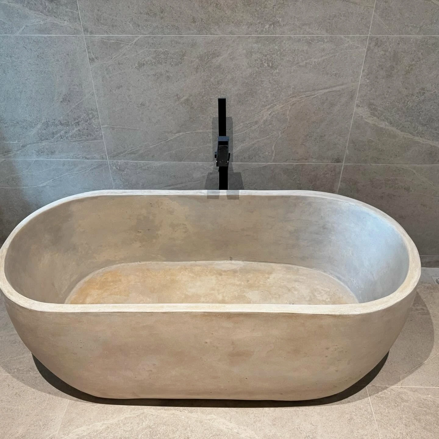 Tina ovalada de concreto beige con grifería negra, en un baño de piso a techo en porcelanato gris