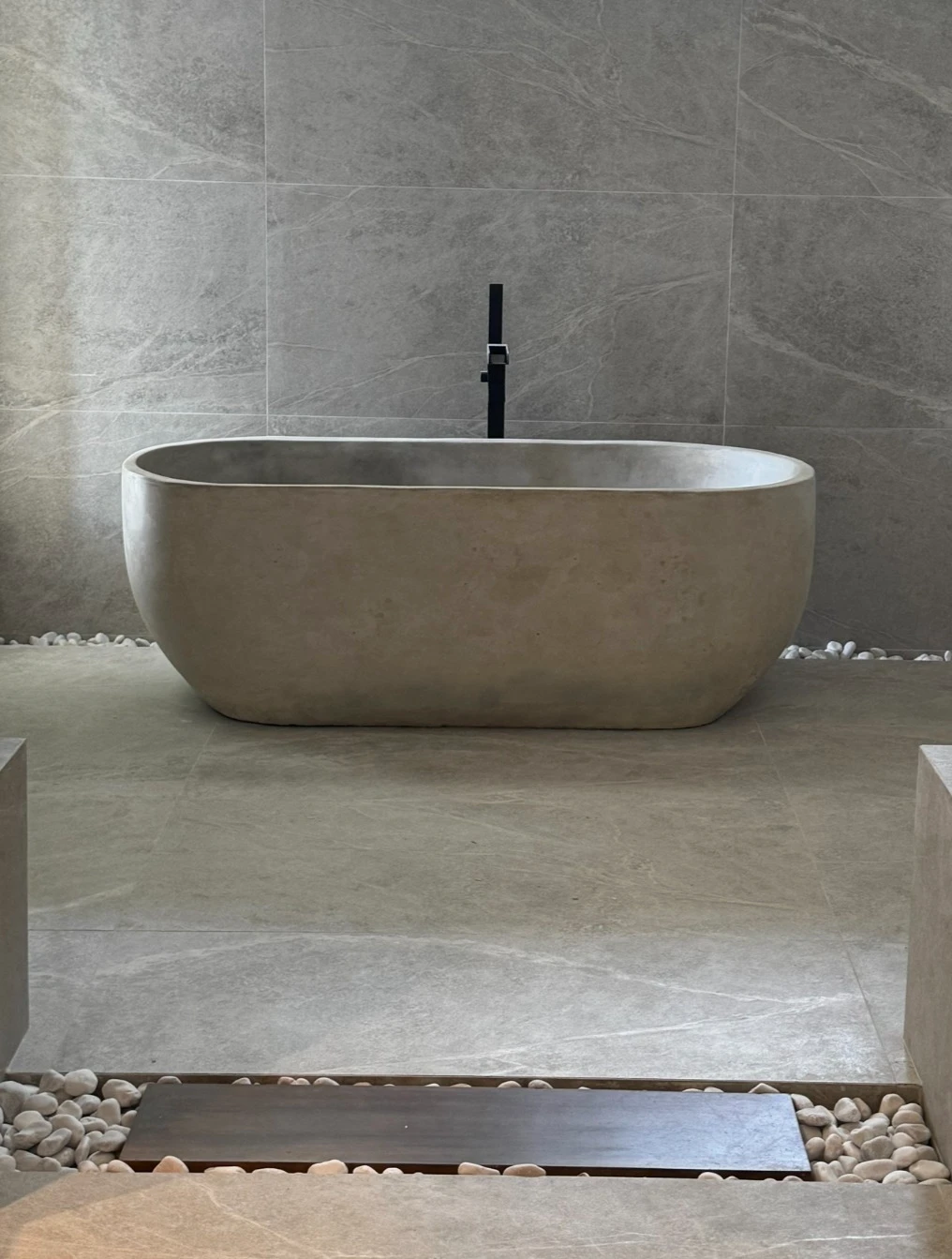 Tina ovalada de concreto claro sobre lecho de piedra blanca, en un baño revestido en porcelanato gris veteado